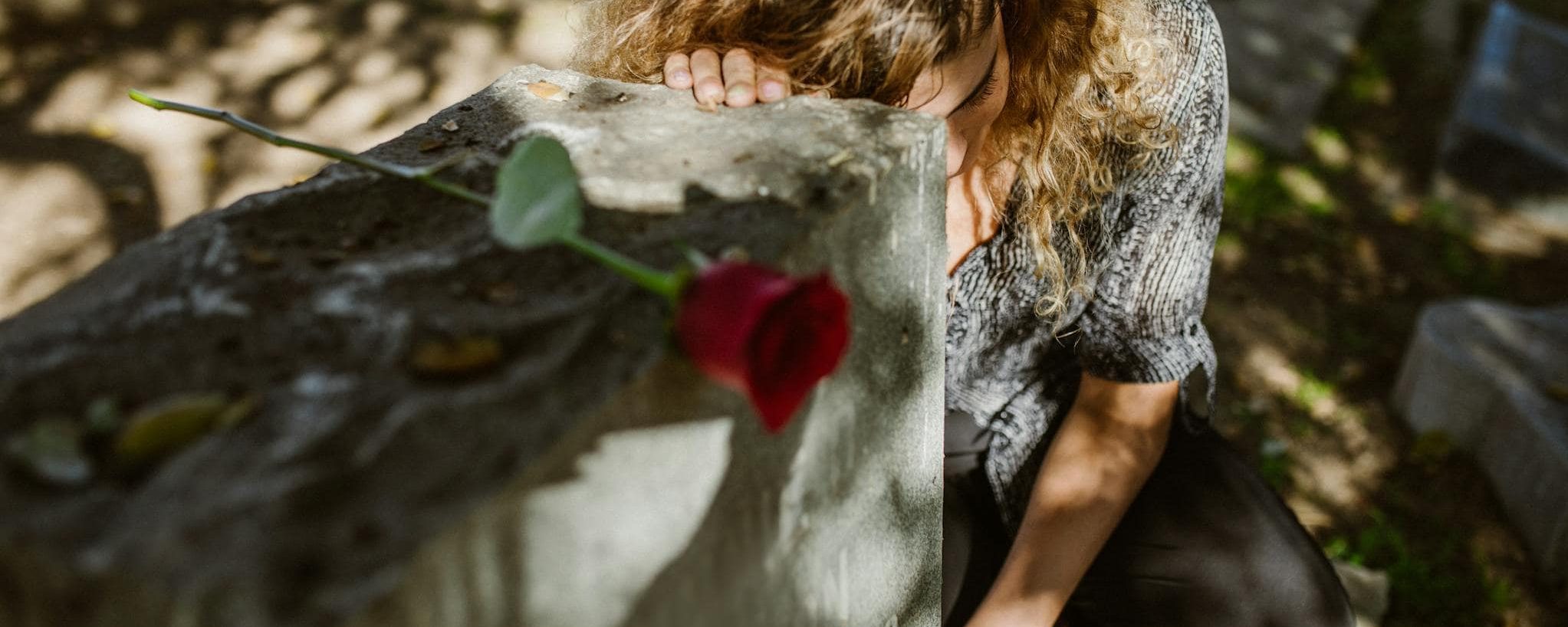 A woman grieves at a cemetery, resting her head on a tombstone with a rose, capturing deep emotion.